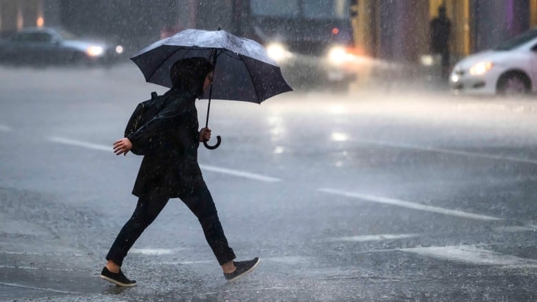 A person wearing all black, including a backpack, crosses an intersection in the rain. They hold an umbrella tilted ahead of them and raindrops patter around their feet.