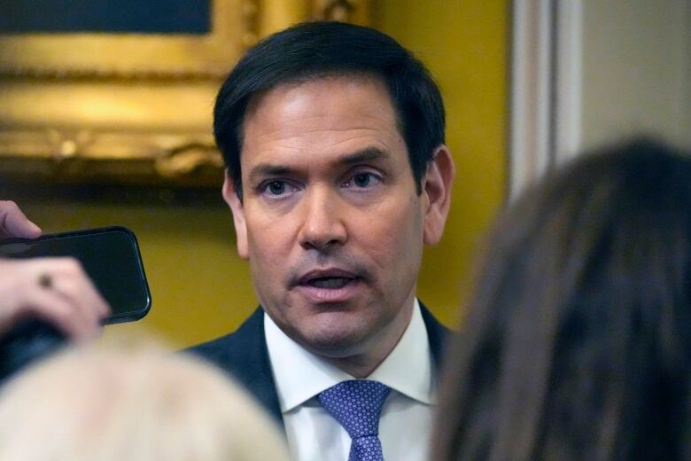 A cleanshaven, dark haired man in a suit and tie is shown in closeup speaking to reporters.