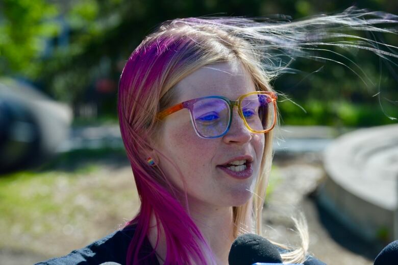 A woman with pink and yellow hair speaks into a microphone. She is wearing glasses with rainbow-coloured frames.