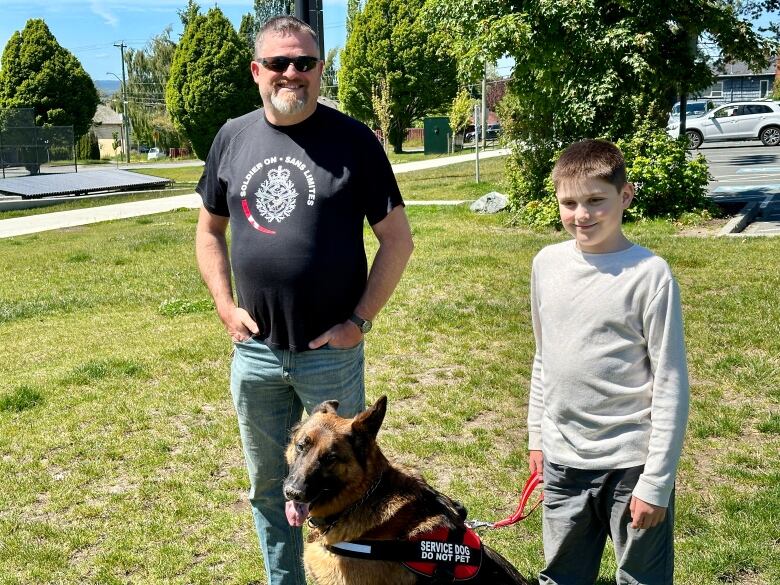 A grown man and a boy stand with a Germn shepherd dog. The dog wears a vest that says 