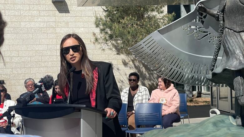 A woman with long dark hair and sunglasses, wearing a black suit jacket with a red design on the lapel, stands at a podium with some people sitting on chairs behind her.