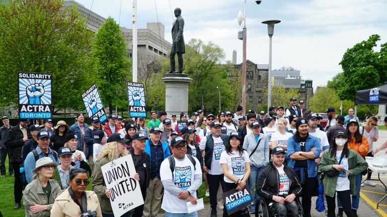 A crowd of people stand on a patio or on a lawn holding protest signs. Many have signs or gear reading 'ACTRA.'