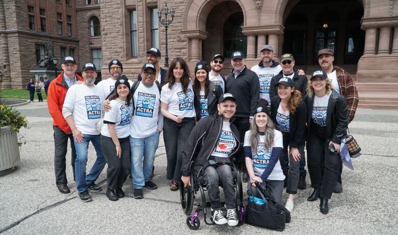 A group of people in ACTRA shirts pose before the staircase of a large old brick building. 