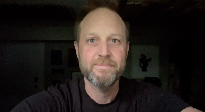 A portrait of a seated person in a black T-shirt against a darkened room. 