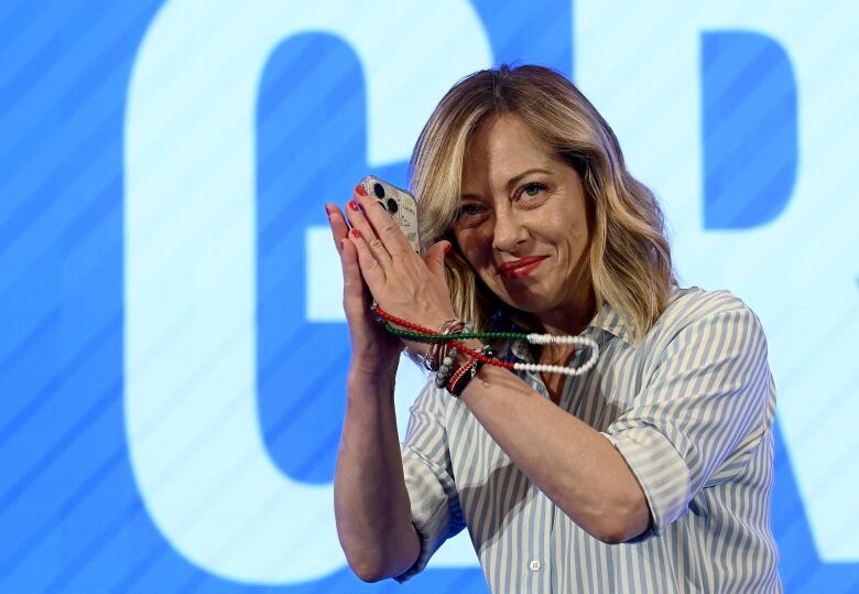 A woman with light hair is shown on a stage with a smile, clasping her hands in a gesture of thanks to an audience.