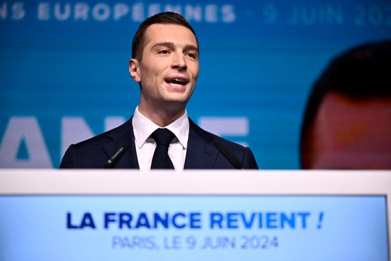 A cleanshaven young man in a suit and tie is shown speaking from a podium that's adorned with the phrase La France Revient.