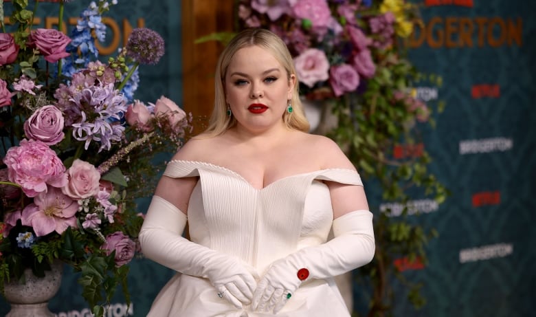 A woman in a white dress and red lipstick poses in front of a floral arrangement