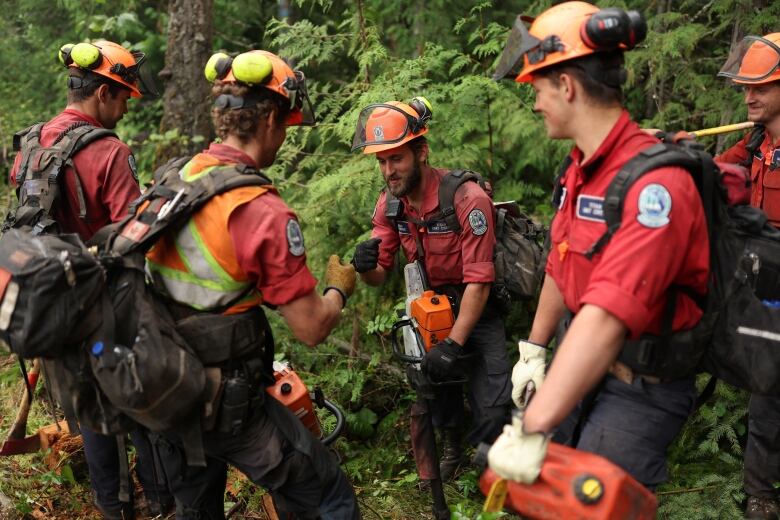 Five men in red shirts and orange helmets smile as they stand together in the woods. The two mid-frame stand with their right hands pointed toward each other in a fist. 