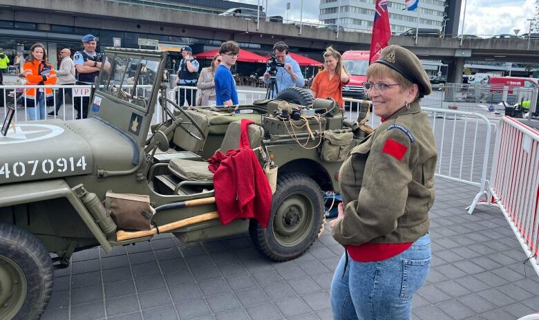 Woman in old-style army jacket and beret in front of a green jeep. 