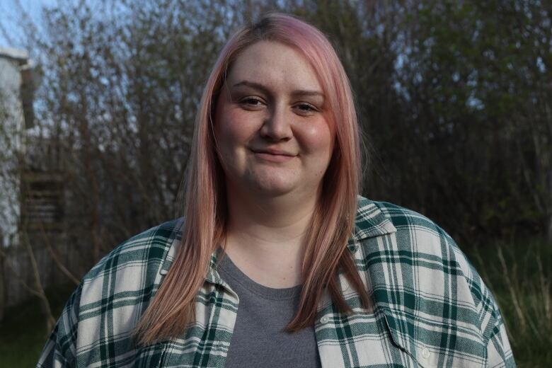 A woman with pink hair and a green shirt standing near some trees.