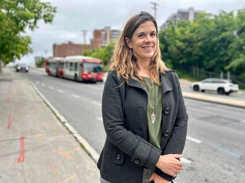 A woman stands on a streetscape looking into the camera, with a city bus passing behind her.