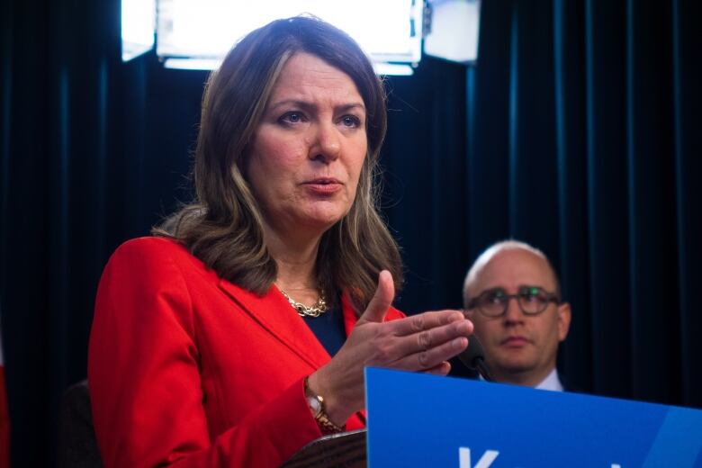 A white woman with brown hair and tinges of blonde speaks at a podium with blue markings.