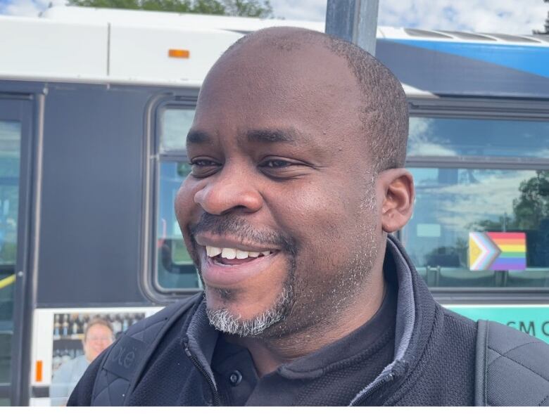 A man with short hair and a neatly trimmed mustache and beard smiles in front of a bus.