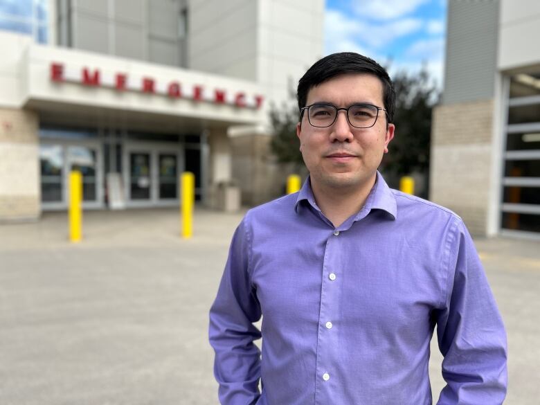 Man with dark hair and glasses wearing a purple shirt stands in front of the emergency department entrance. 