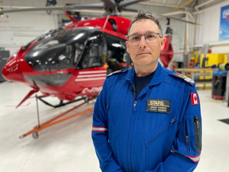 Man with grey hair and glasses stands wearing a blue flight suit in front of a STARS helicopter at the Winnipeg base. 