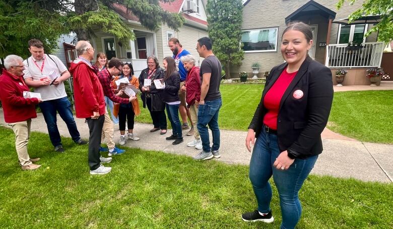 A woman in a blazer and jeans stands on a boulevard, with a group of volunteers in the background.