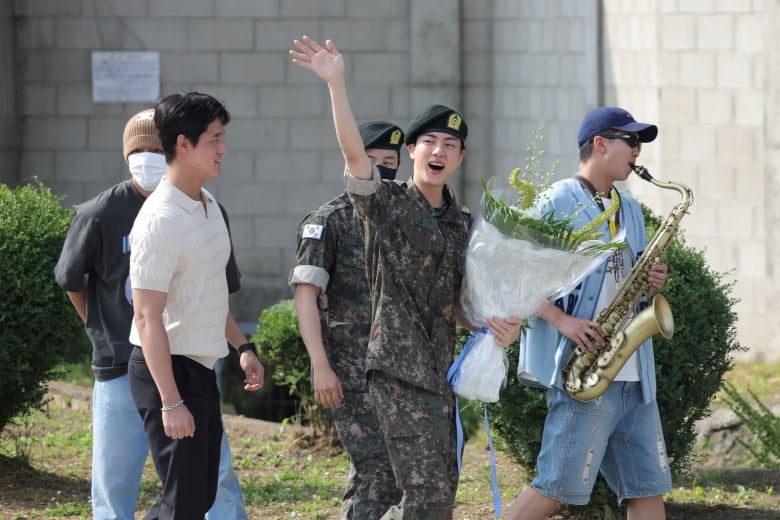 A young man in a military uniform and beret waves as he walks. Another young man beside plays the saxophone.