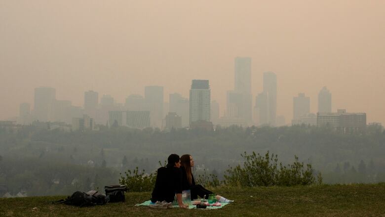 A young couple sit on the grass over looking the city of Edmonton skyline that is covered in smoke from wildfires.