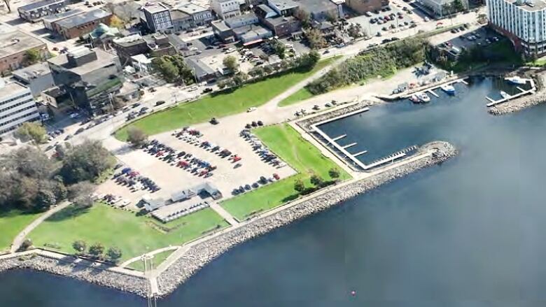 An aerial photo of a city waterfront with blue water, green space, a gravel parking lot and a marina where a few boats are docked.