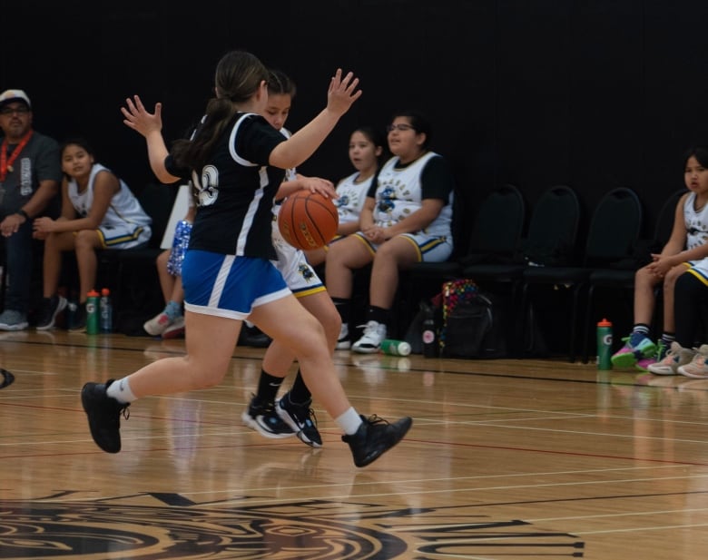 An Indigenous girl dribbles a basketball down a court while another attempts to block her. 