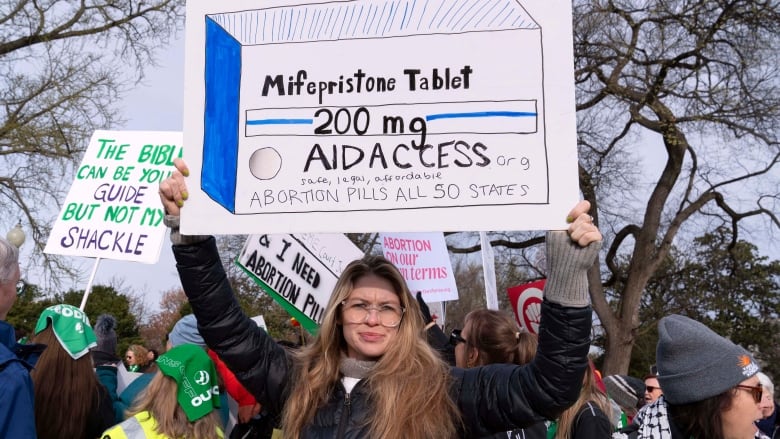 A woman with long hair and glasses holds a sign over her head as other people wearing coats and hats are shown outdoors at a demonstration. 