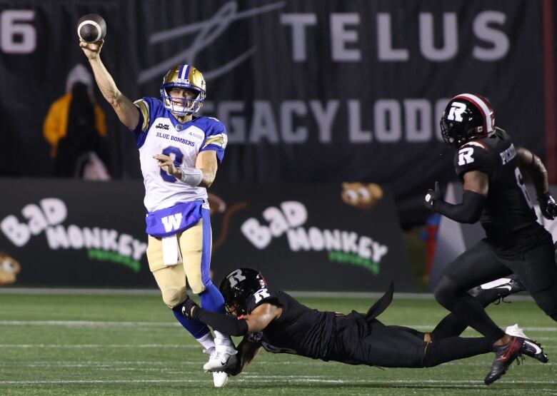 A football player in black dives at the ankles of a quarterback in white, just as he releases the football.