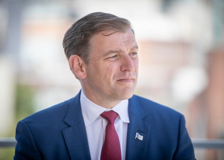 A man in a blue blazer with a white shirt and red tie looks to his right side. He is wearing a lapel pin of the Newfoundland and Labrador flag. 