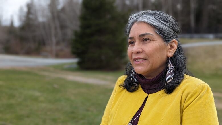 A woman with shoulder-length hair is wearing dangly earrings and a turtle-neck. She is in a park with trees behind her.