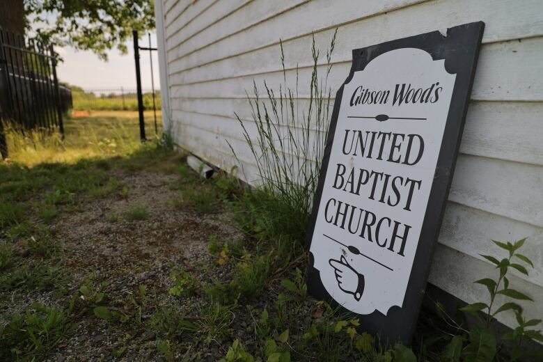 A sign leans against a white building and says, Gibson Woods United Baptist Church.