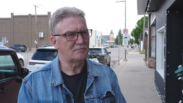 A man with short grey hair, black-rimmed glasses, and wearing a blue jean jacket looks forward.