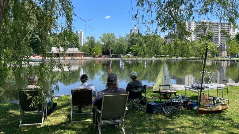 People in lawn chairs face away from camera to look at little remote control boats on small lake