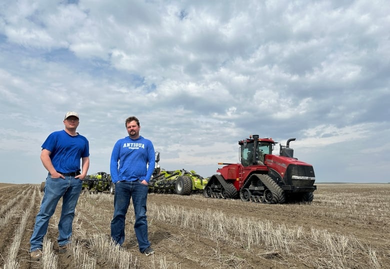 Two men stand in front of a seeder and tractor in a field.