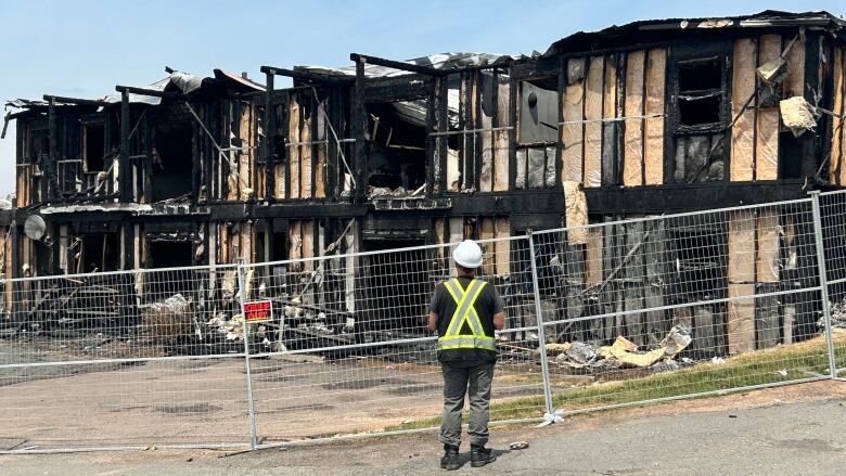 A man in a hard hat and a safety vest stands in front of a chain link fence with a burned down two-storey apartment complex in the background.