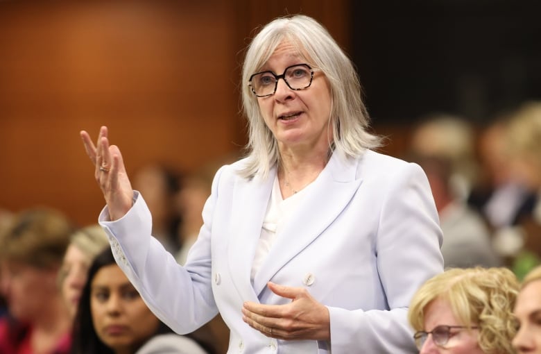 A woman in a white suit speaks with her arms raised at a meeting.