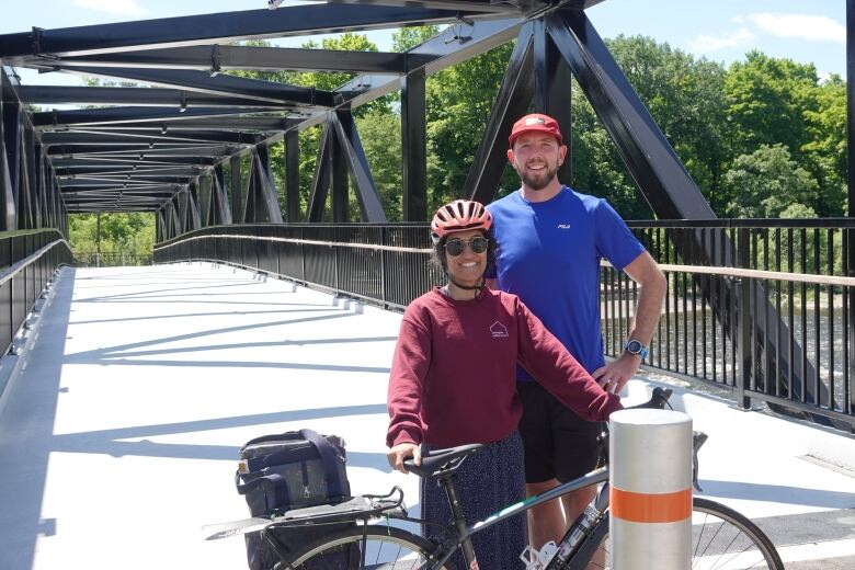 A man and a woman smile for a photo with a bicycle in front of a footbridge.