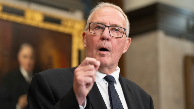 A man in a suit with Grey Hair holds his hand up as he speaks to reporters from the foyer of the House of Commons. 