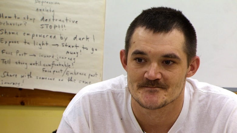 A man wearing a white t-shirt sits in front of a whiteboard