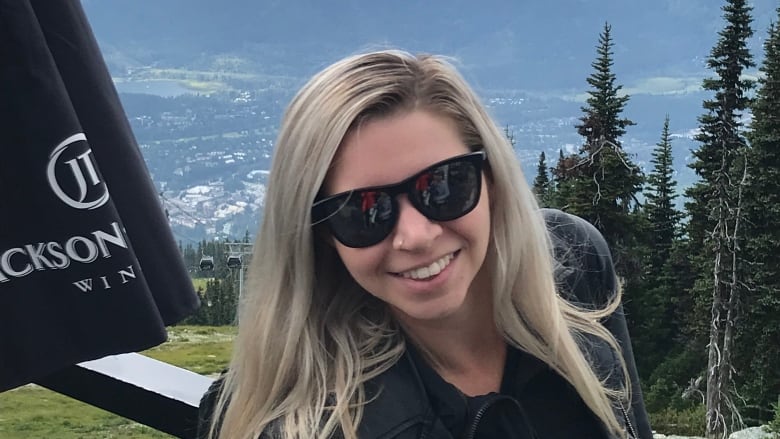 A white woman with long blond hair wearing a black jacket and black sunglasses stands in front of a mountain vista.