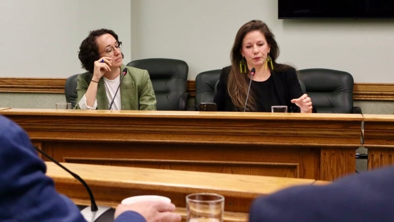 A woman wearing a green jacket and a white shirt sits next to a woman in a black jacket and black shirt. Both women are sitting at a wooden desk. 