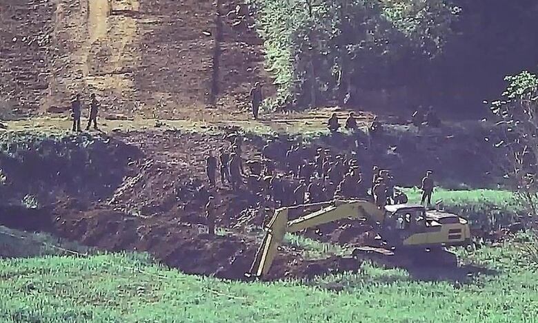 A grainy still from a video shows workers from a distance who appear to be wearing military uniforms while standing on a grassy area near a crane.