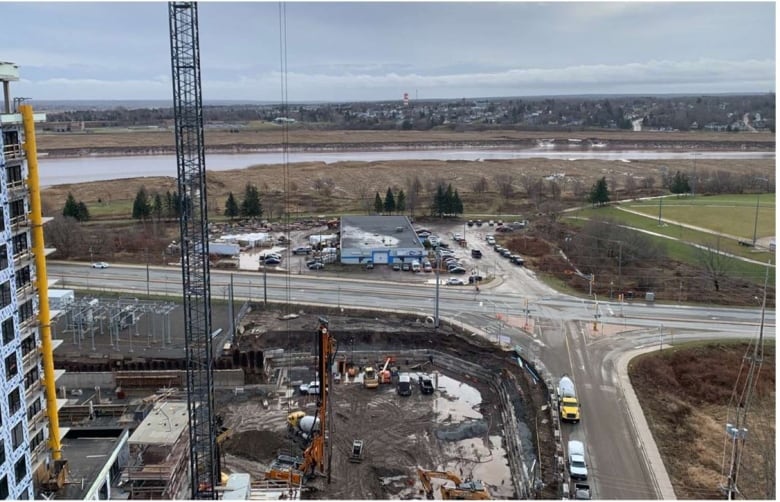 An elevated view showing a construction site, tower crane, various vehicles and equipment, streets and the riverfront in the background.