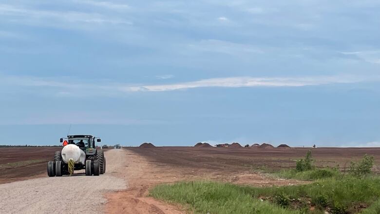 A tanker truck drives down a gravel road toward a peat bog. Small mounds of peat moss are seen on the horizon.