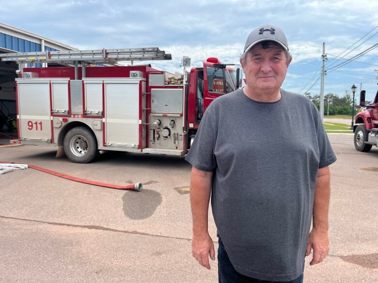 A man wearing a grey baseball cap and t-shirt stands in front of a red fire truck with a hose on the ground.