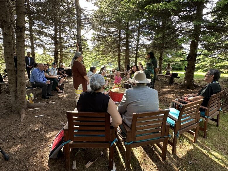 A group of people sit on wooden chairs outside in a forest. 