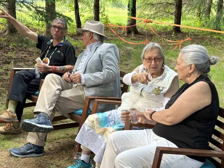 Two men and two women sit in wooden chairs in a forest.