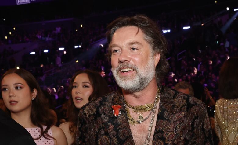 A man with brown hair and a grey-white beard looks off camera at an awards ceremony where he's wearing a fancy jacket with his bare chest adorned with necklaces underneath. 