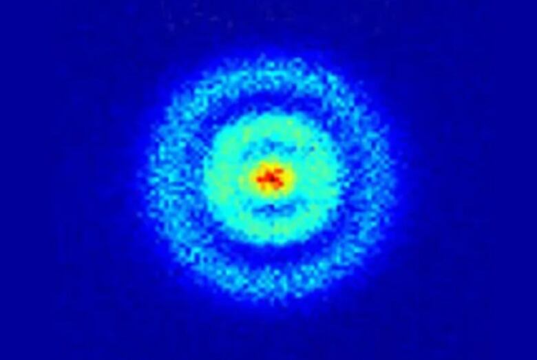 On a royal blue background, we see a fuzzy circular object in the centre with a bright red dense nucleus and aqua blue rings around it to show where the electron wave / particles are located. 