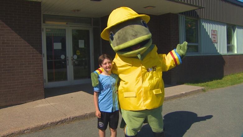 A smiling boy wearing a Toronto Blue Jays jersey stands next to a large cod fish mascot wearing a yellow rain coat and sou'wester hat.