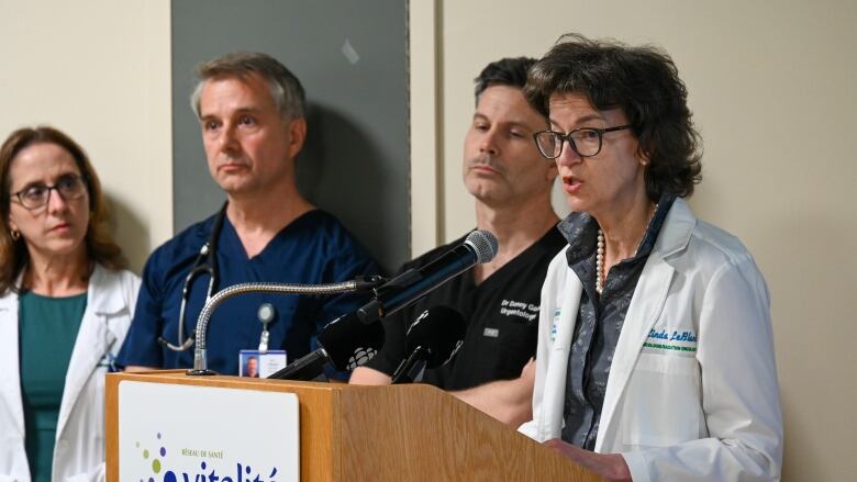 A woman in a white lab coat speaks at a podium next to three other people.