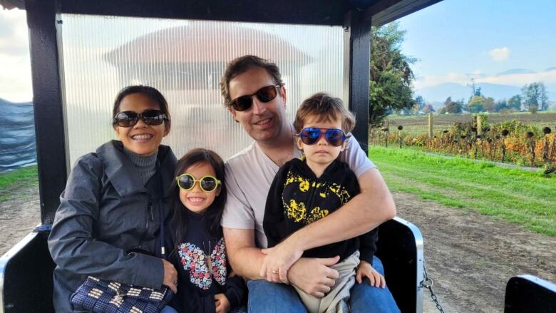 A family of four sits on a cart with a farm behind them.